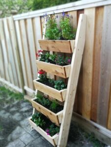 Wooden multi-tier vertical planter leaning against a fence with several planting boxes filled with flowers and herbs