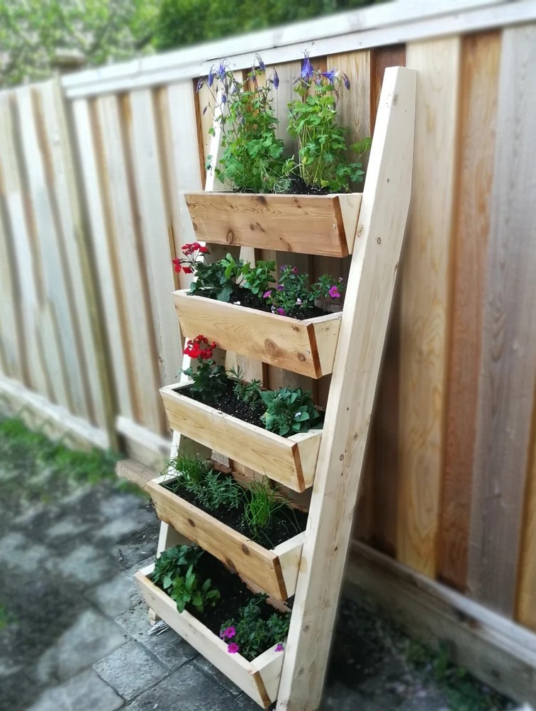 Wooden multi-tier vertical planter leaning against a fence with several planting boxes filled with flowers and herbs